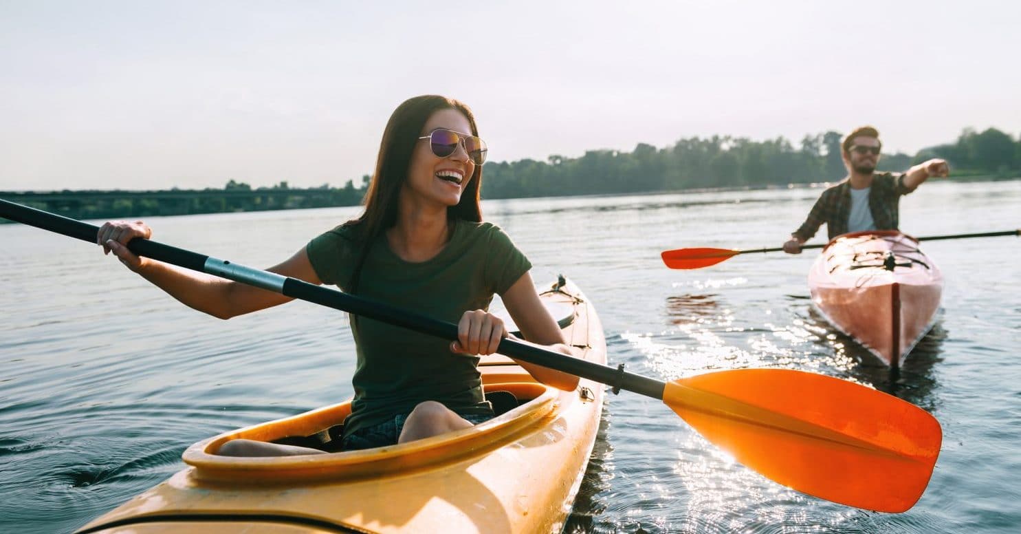 A woman paddles a yellow kayak on a lake, smiling, while a man in a pink kayak gestures nearby. A woman paddles a yellow kayak on a lake, smiling, while a man in a pink kayak gestures nearby.