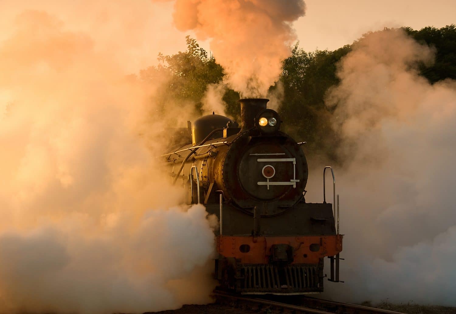 A vintage steam locomotive emerges from a cloud of steam at sunset.