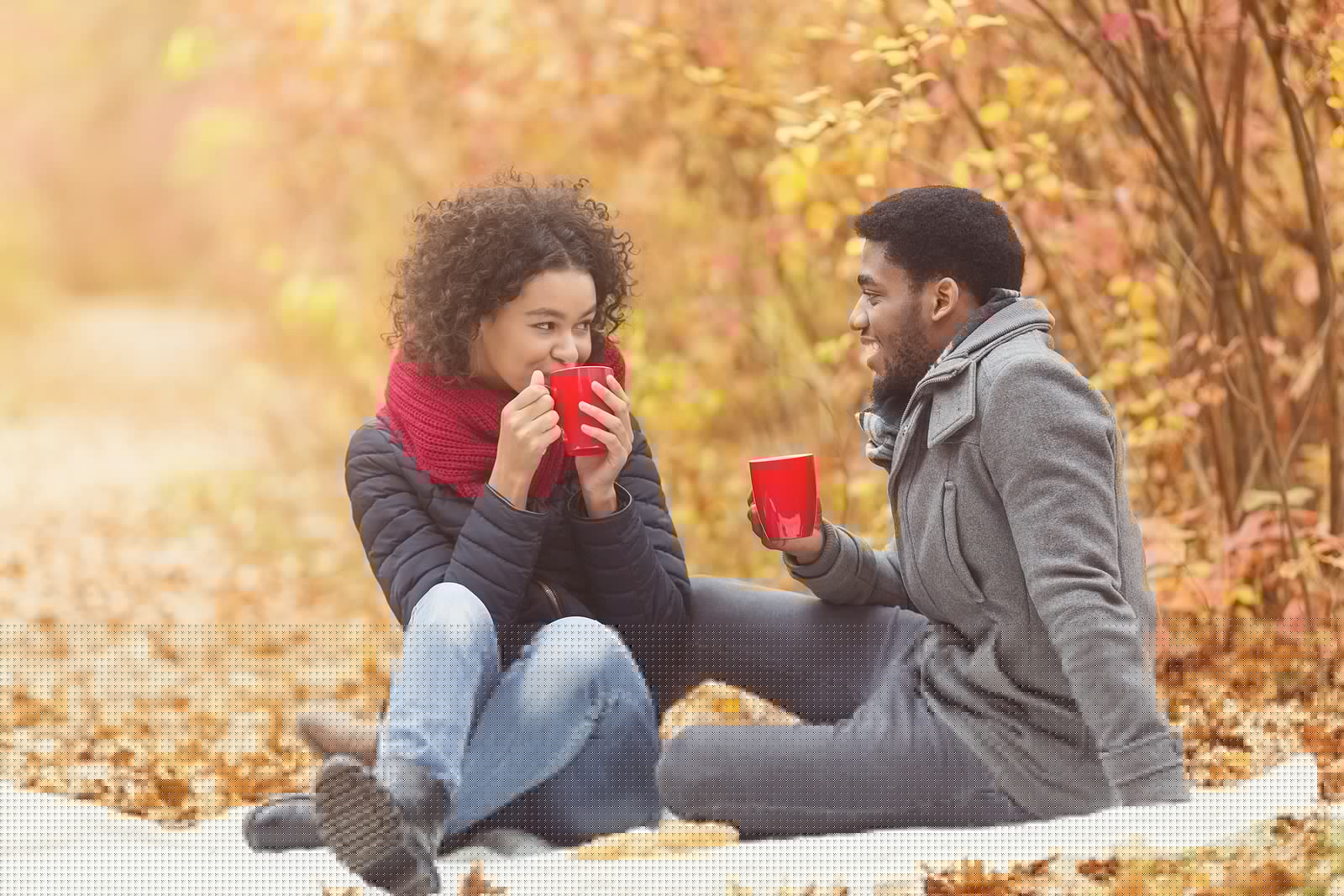 A couple sits on a blanket in a fall setting, enjoying warm drinks and smiling at each other.