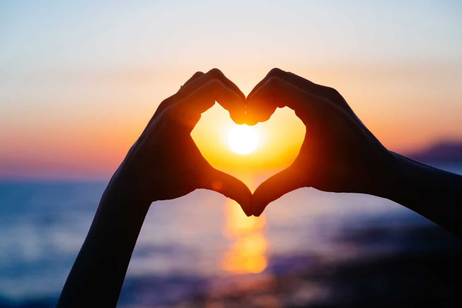 Hands forming a heart shape against a sunset over the ocean.