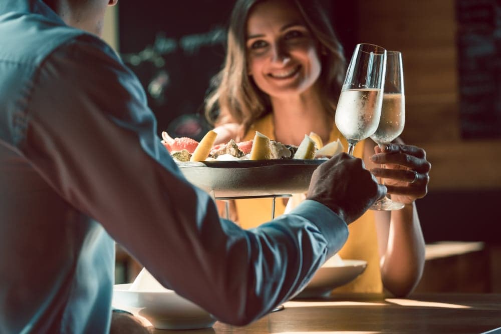 A man serving a seafood platter to a smiling woman who is raising a glass of sparkling wine.