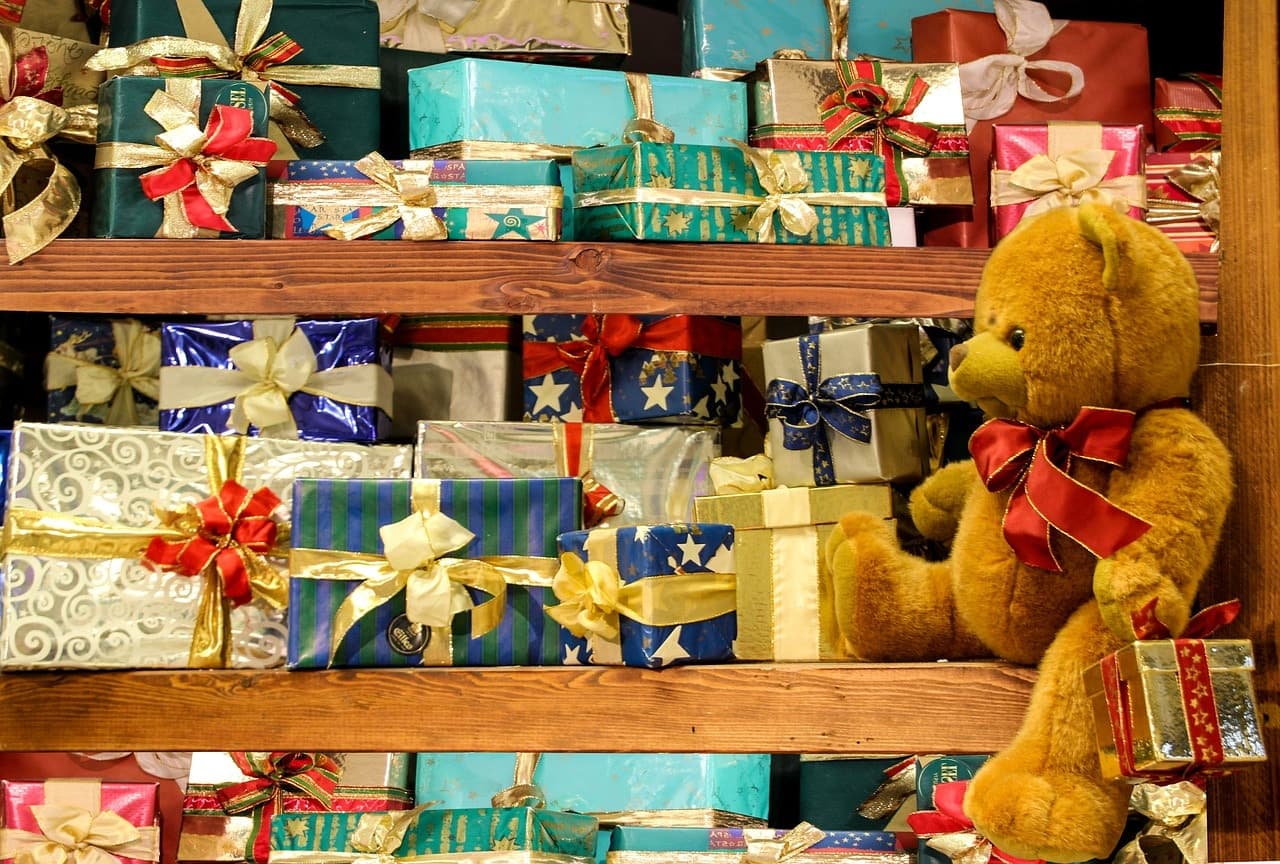 A stuffed teddy bear sits beside a colorful display of wrapped gifts on wooden shelves.