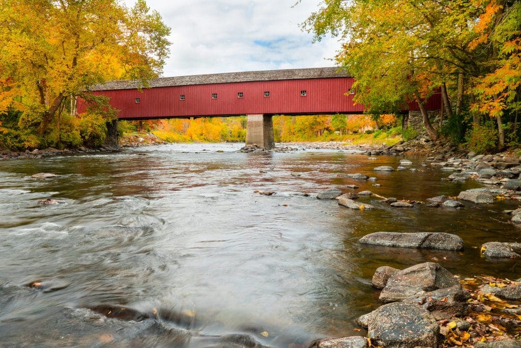 A red covered bridge spans a tranquil river surrounded by vibrant autumn foliage.