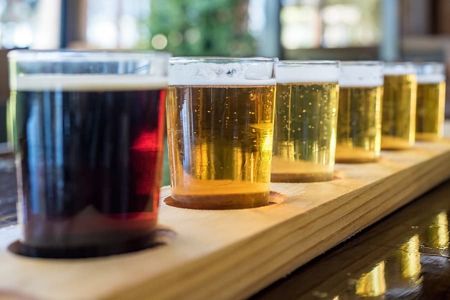 A wooden flight board holds six small glasses of various beers, ranging from dark to light.