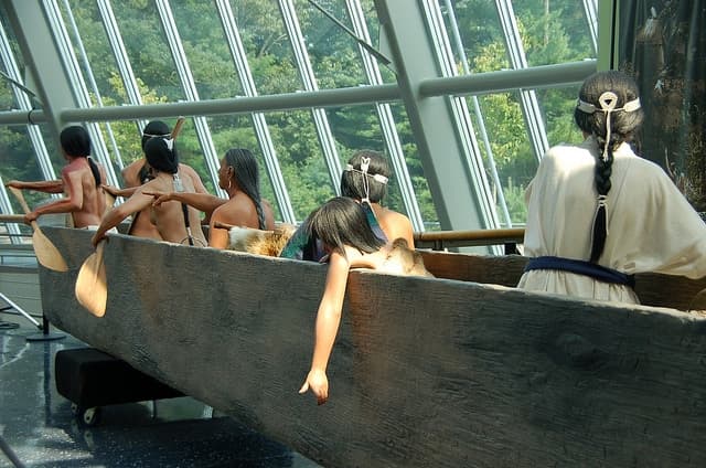 A group of life-sized figures in traditional clothing is paddling a canoe indoors, surrounded by large windows.