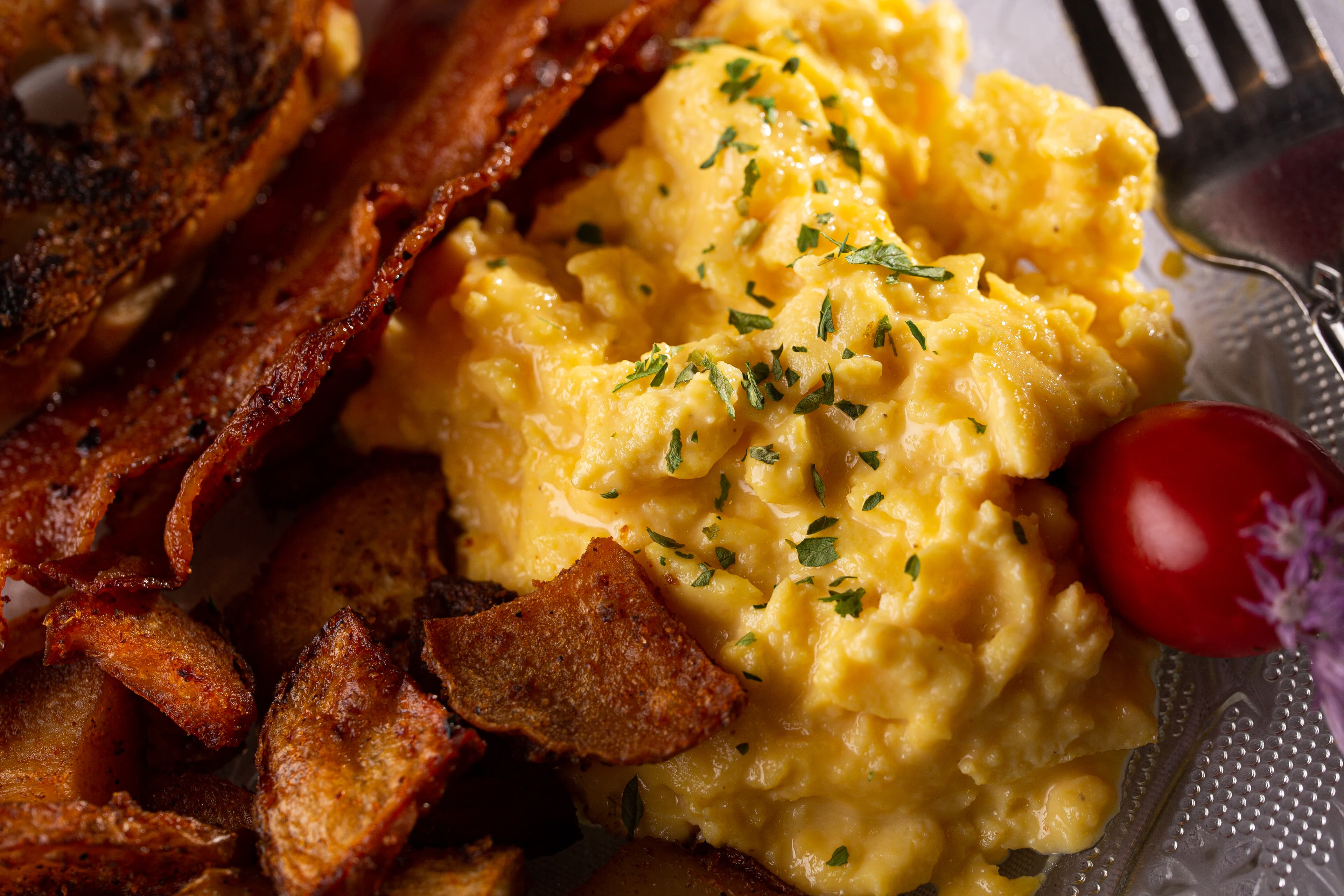 A plate of fluffy scrambled eggs with crispy bacon, golden potatoes, and a cherry tomato.
