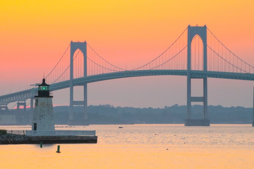 A lighthouse stands by the water, with a bridge spanning the horizon against a colorful sunset.