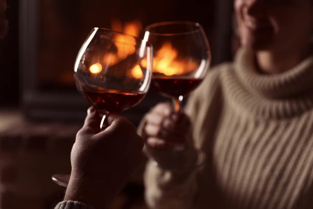 Two people clink wine glasses together in front of a cozy fireplace.