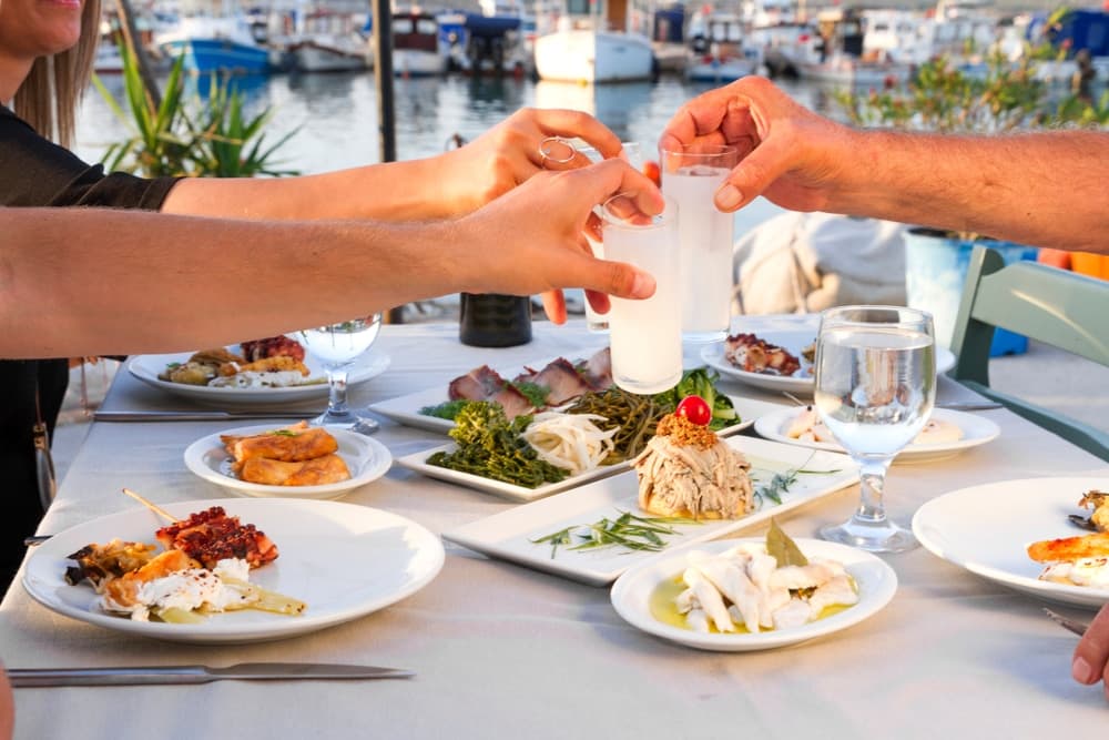 Two people toast drinks over a table filled with various dishes near a waterfront setting.