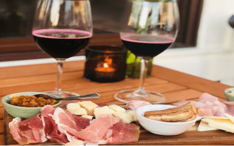 A wooden platter with assorted meats, cheeses, and a dipping sauce, accompanied by two glasses of red wine and a candle on a table.