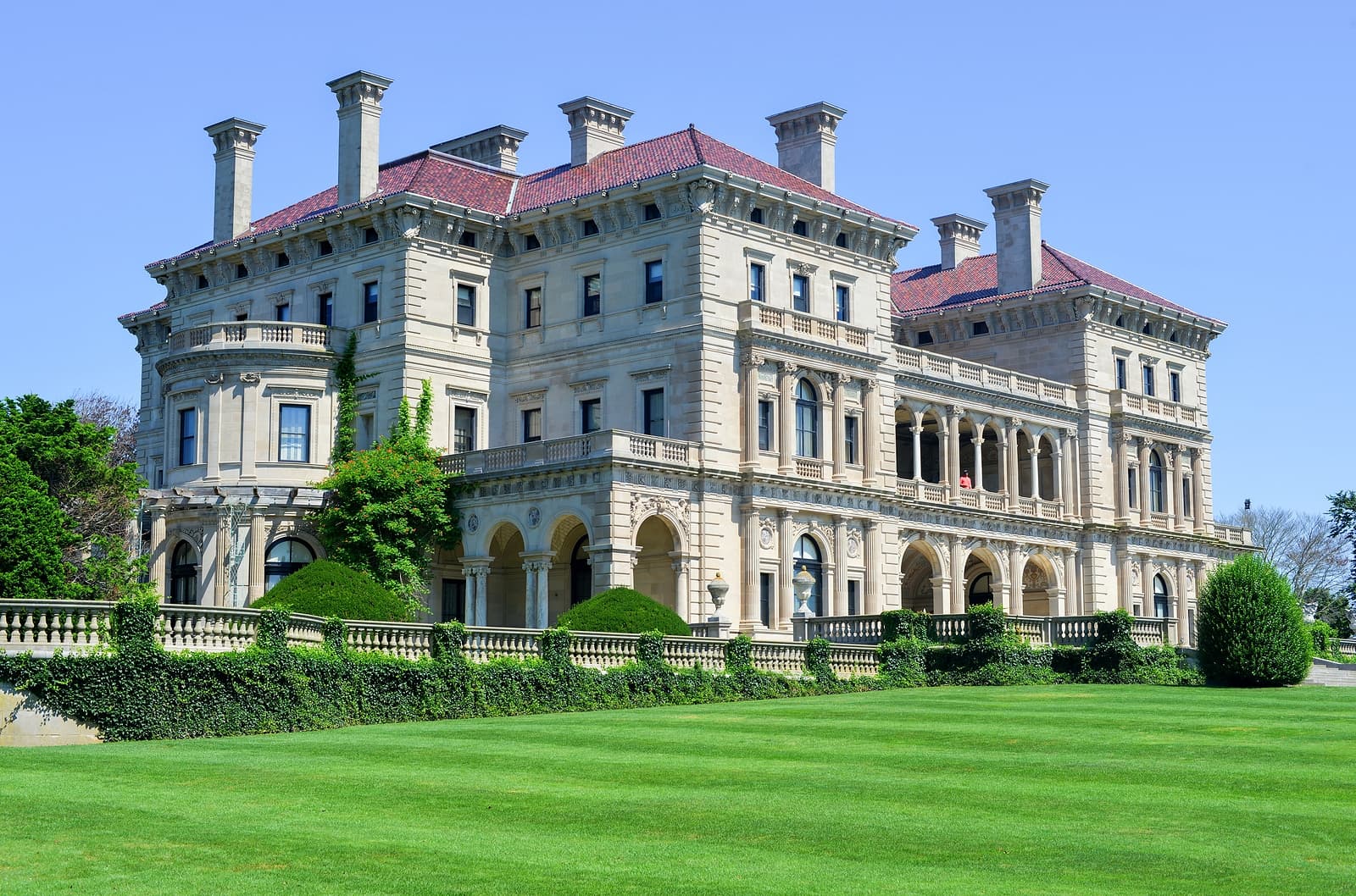 A grand stone mansion with a red-tiled roof, surrounded by a manicured lawn and greenery.