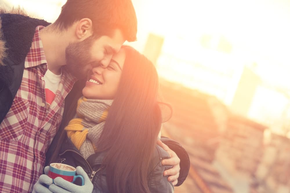A couple smiles affectionately at each other while sharing a warm drink outdoors in a sunlit setting.