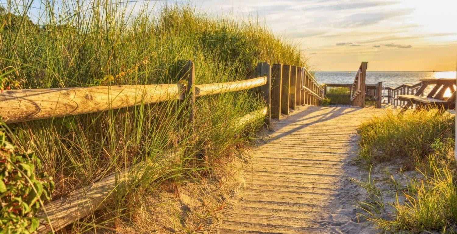A wooden boardwalk lined with tall grass leads to the beach at sunset.