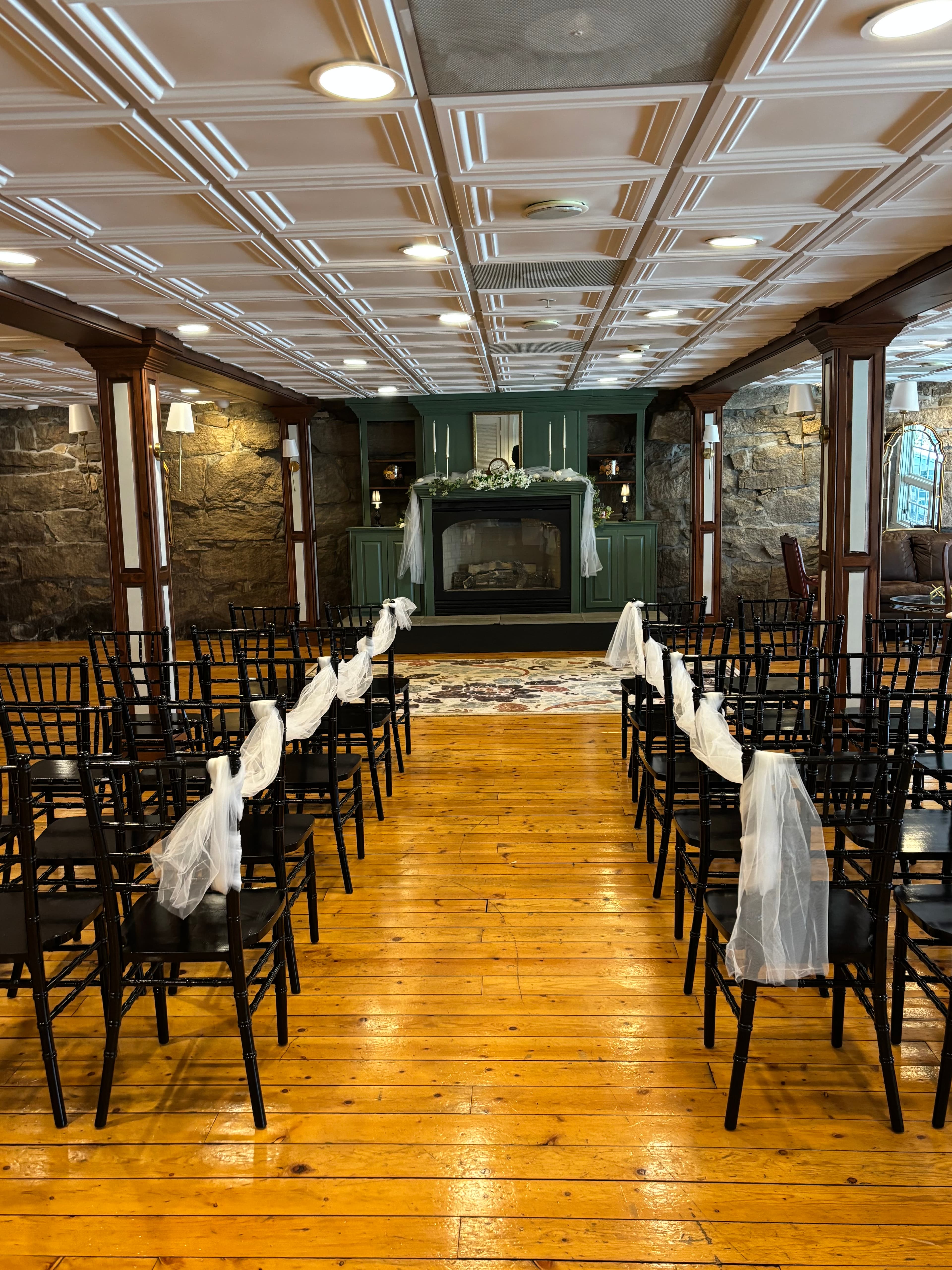 A wedding ceremony setup featuring black chairs adorned with white fabric, facing a decorated fireplace in a warmly lit venue.