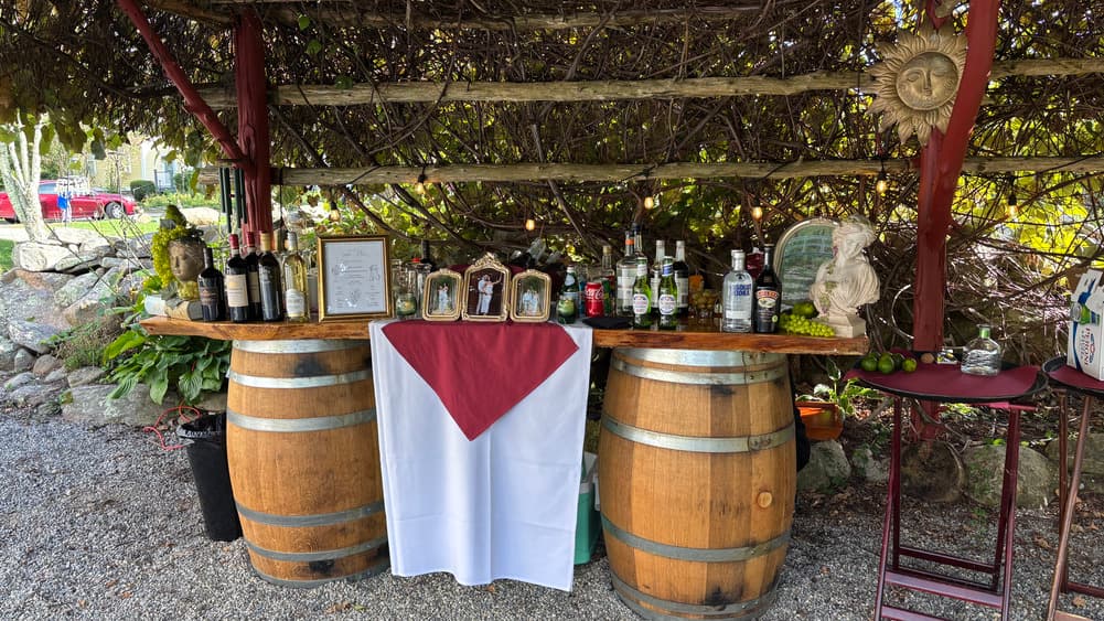A rustic bar setup made from wooden barrels, adorned with a white tablecloth featuring a red triangular accent, displaying various bottles and framed pictures under a vine-covered structure.