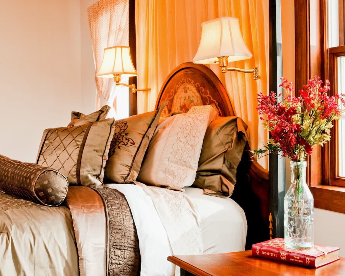 A cozy bedroom featuring a bed with decorative pillows, bedside lamps, and a vase of flowers on a wooden nightstand.