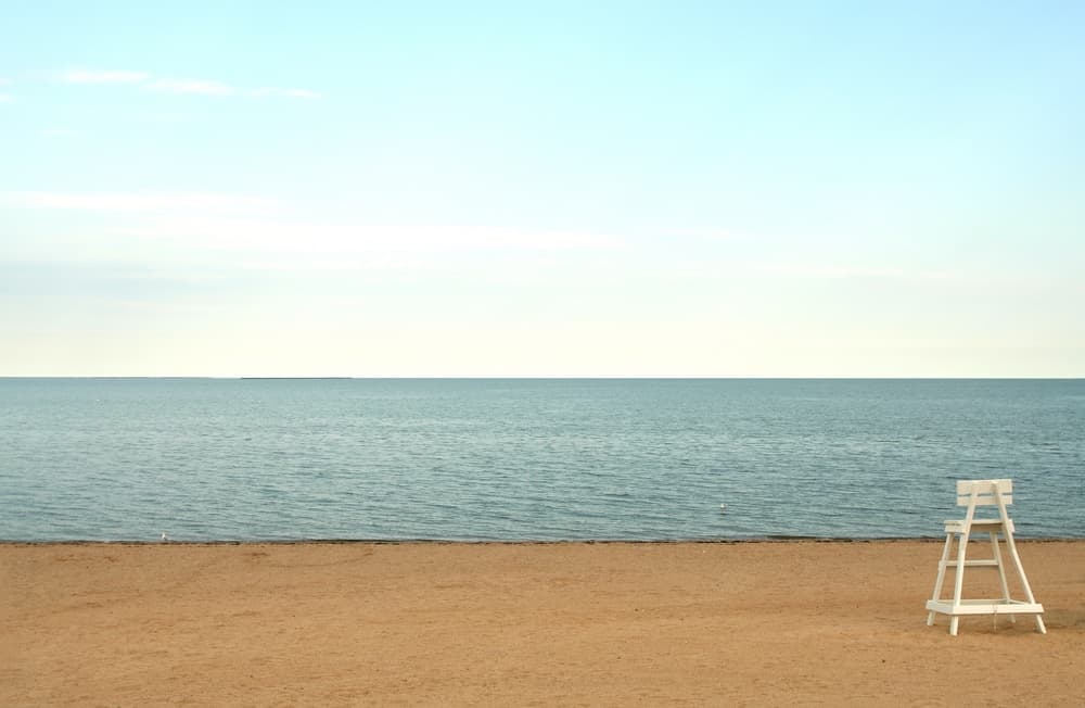 A lifeguard chair stands on a sandy beach facing a calm sea under a clear sky.