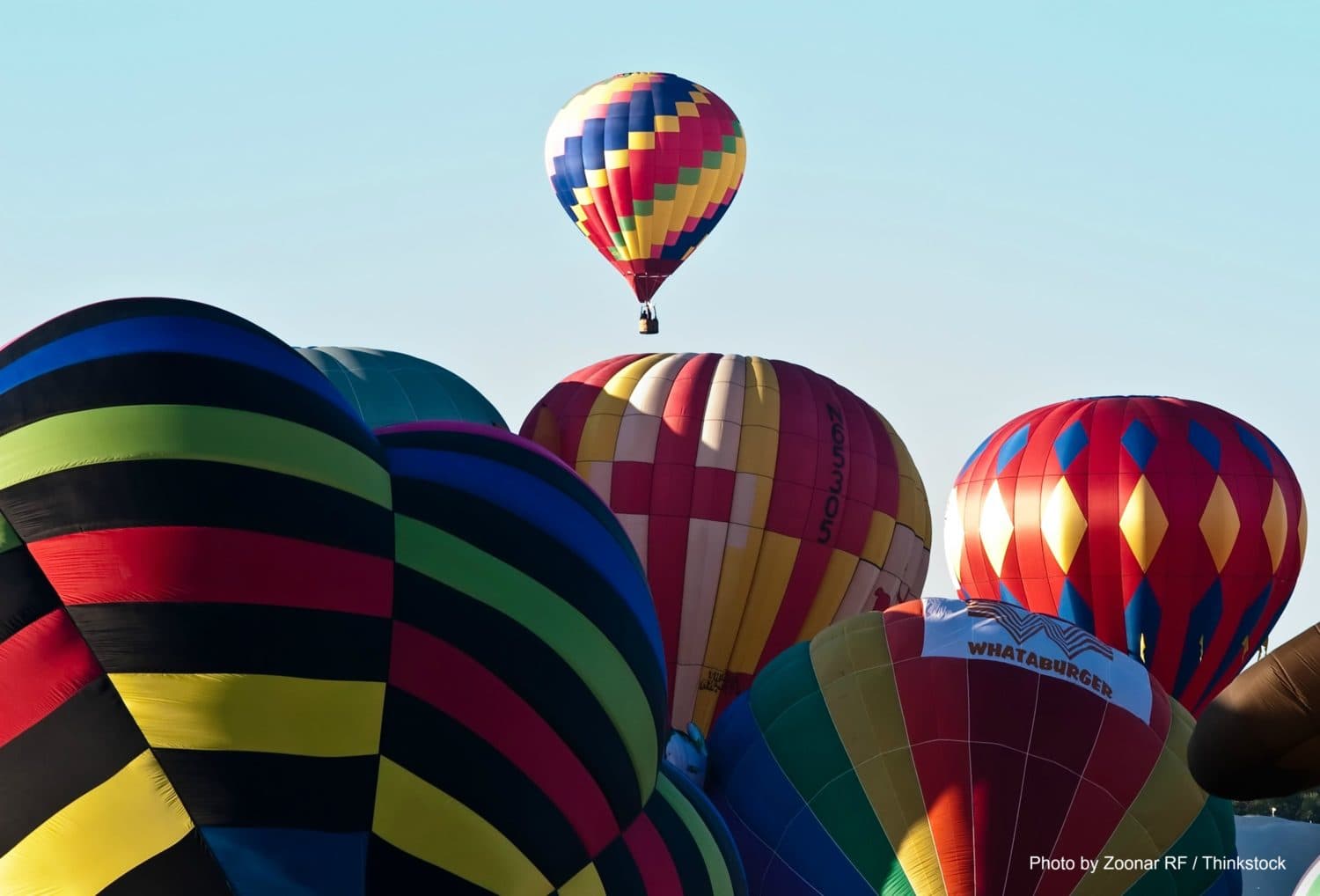 A colorful array of hot air balloons with one balloon rising above the others against a clear sky.