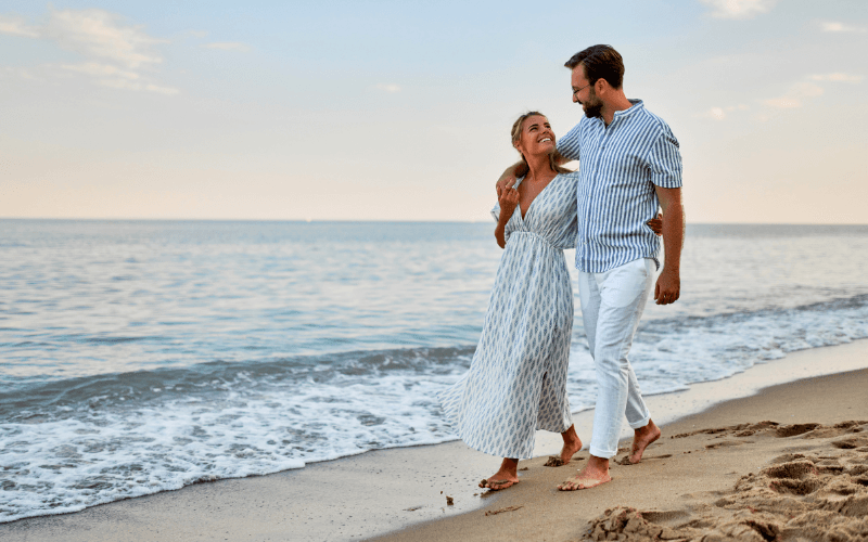 A couple walks hand in hand along a beach at sunset, smiling at each other.