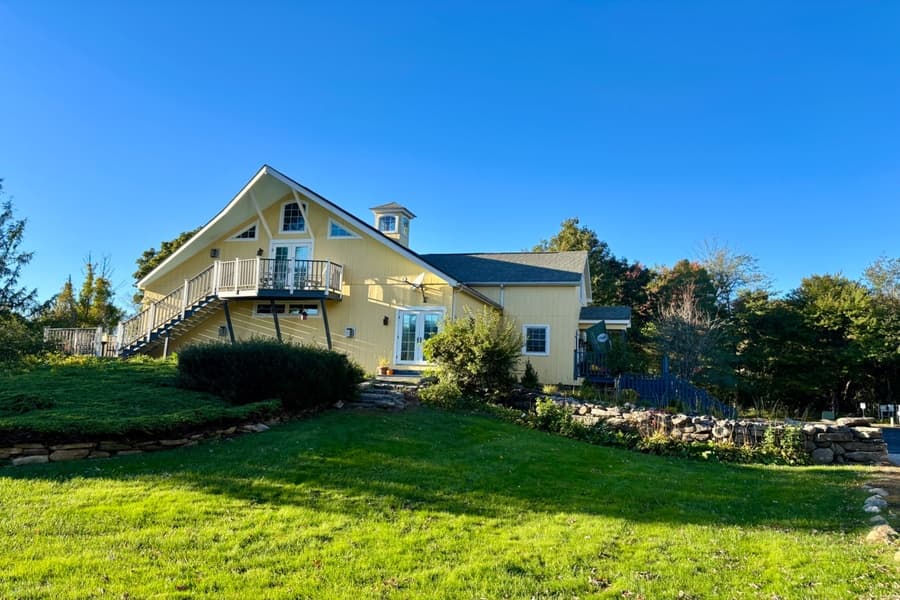 A bright yellow house with a balcony, surrounded by green grass and trees under a clear blue sky.