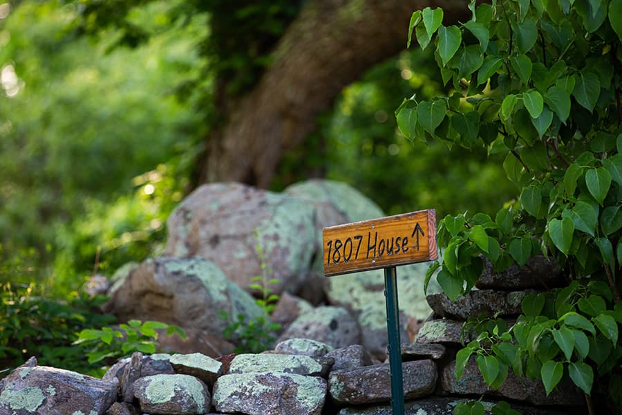 A wooden sign pointing towards "1807 House" is surrounded by greenery and rocks.