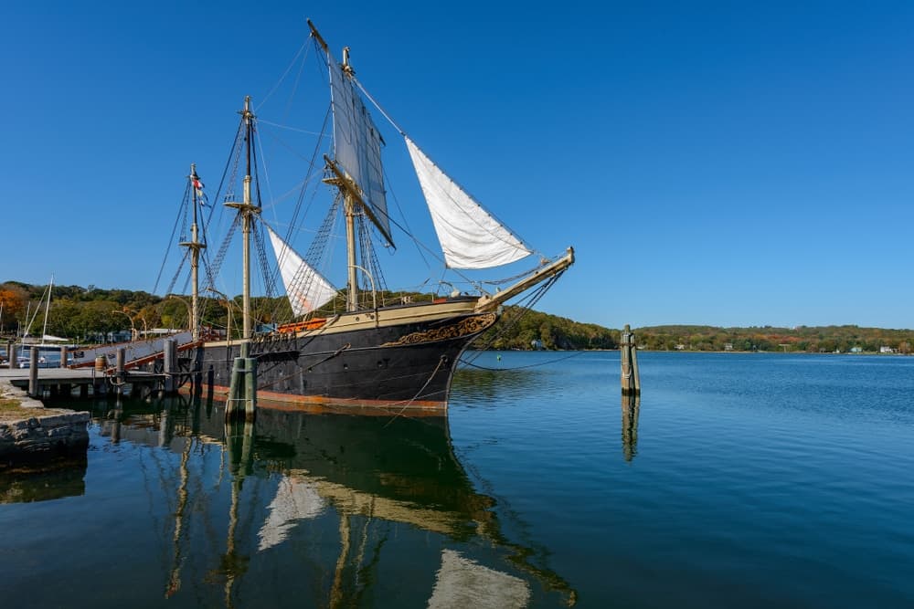 A tall ship with white sails docked by a calm waterfront under a clear blue sky.