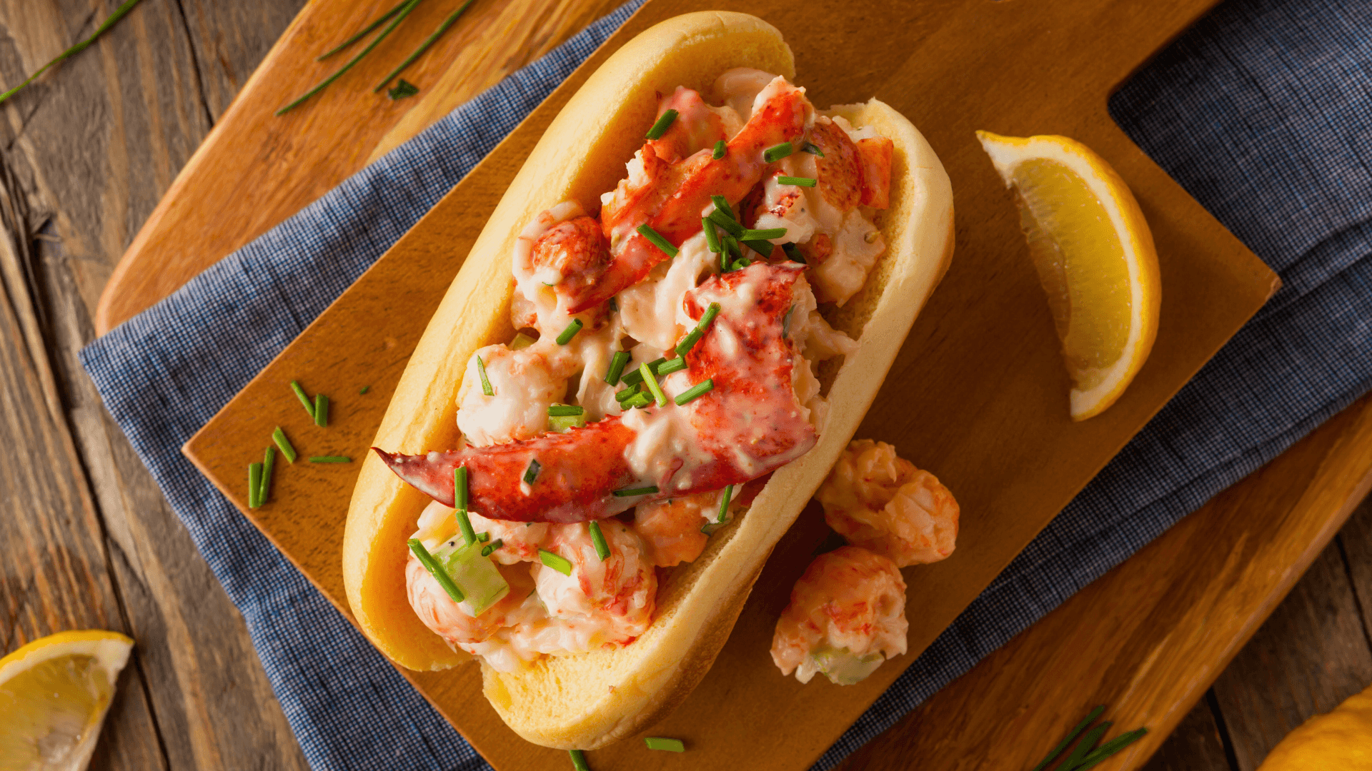 A lobster roll topped with chives, served with lemon wedges on a wooden platter.