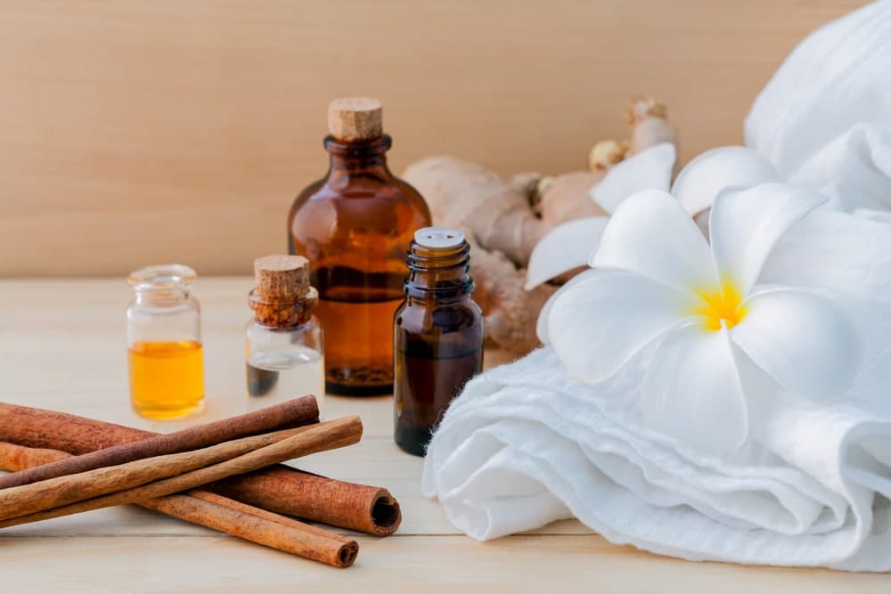 Bottles of essential oils, cinnamon sticks, ginger, and a white flower are arranged on a wooden surface with a towel.