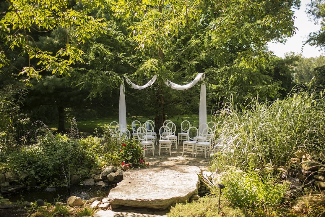 A serene outdoor wedding setup with white chairs and flowing drapes beneath a tree.