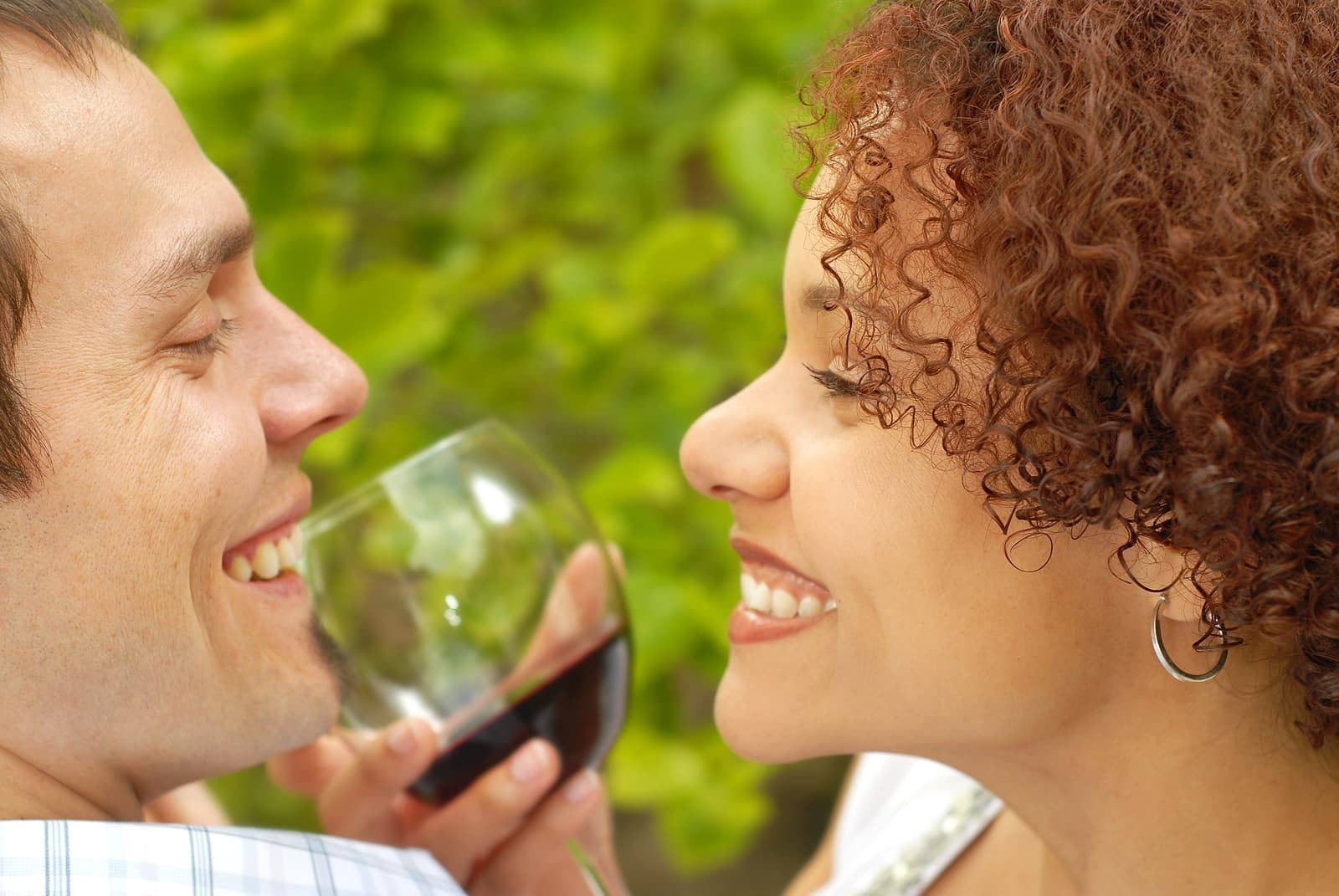 A couple smiles at each other while sharing a glass of wine against a greenery backdrop.