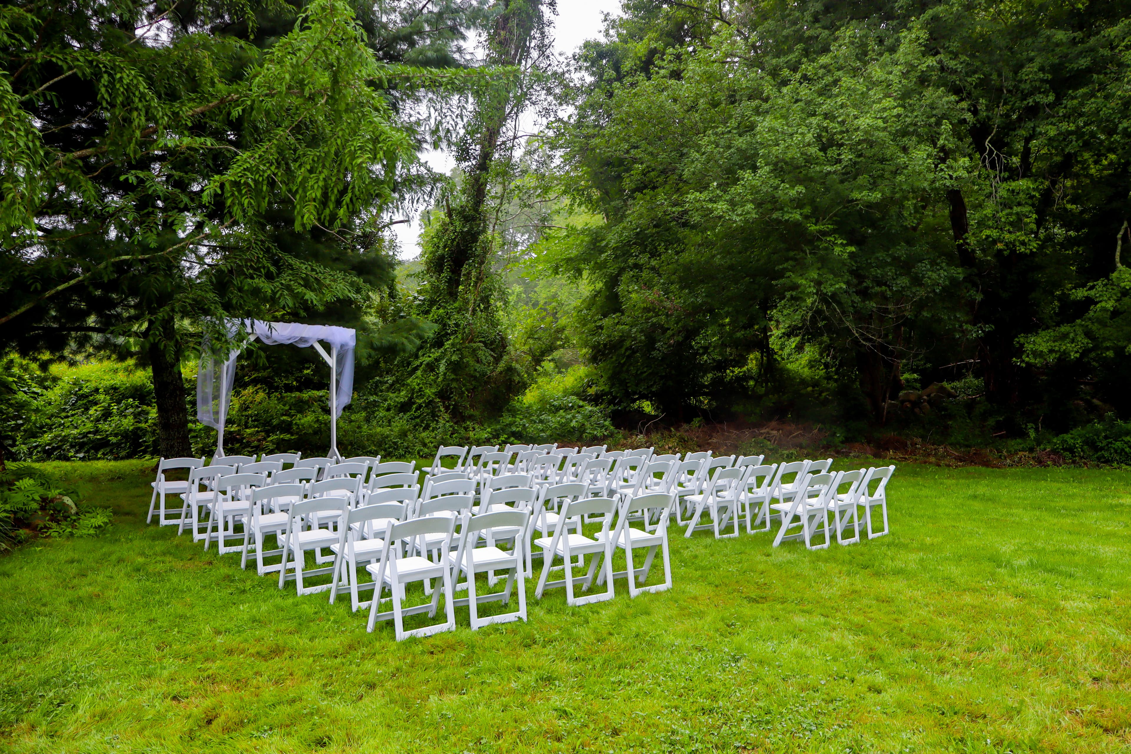 A set of white chairs arranged in rows under a simple canopy in a grassy area surrounded by trees.
