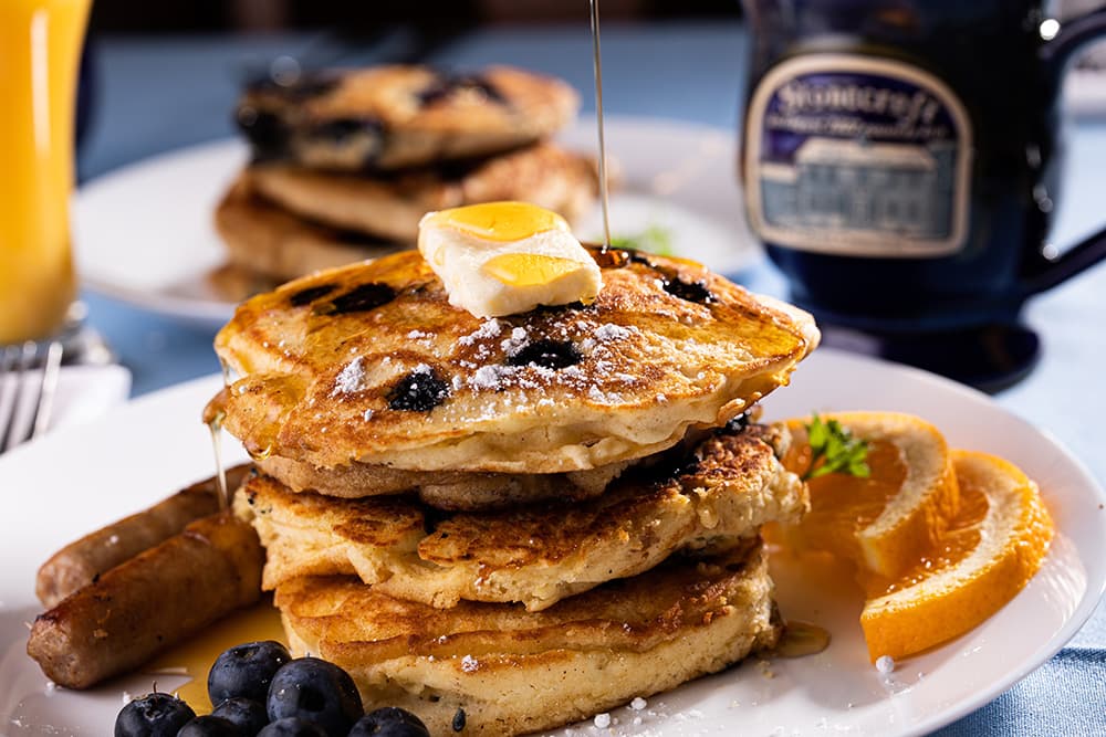 A stack of blueberry pancakes topped with butter and syrup, accompanied by sausage, blueberries, and orange slices.