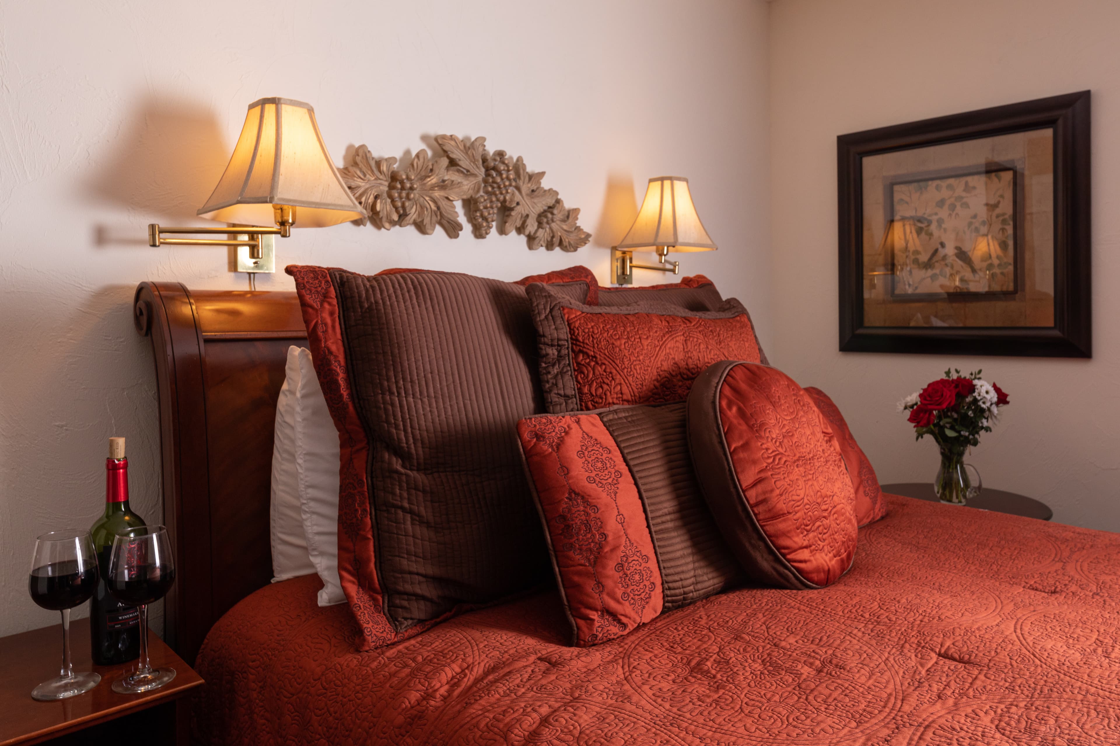A cozy bed adorned with rich, reddish-brown pillows, flanked by wall lamps, a framed picture, and a side table with wine and flowers.