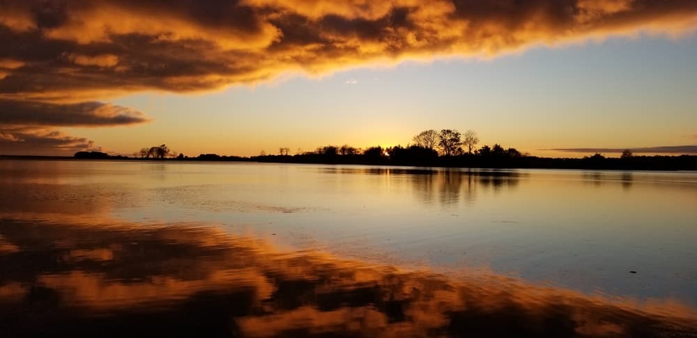 A tranquil sunset over a calm lake, with silhouetted trees and vibrant clouds reflected in the water. A tranquil sunset over a calm lake, with silhouetted trees and vibrant clouds reflected in the water.