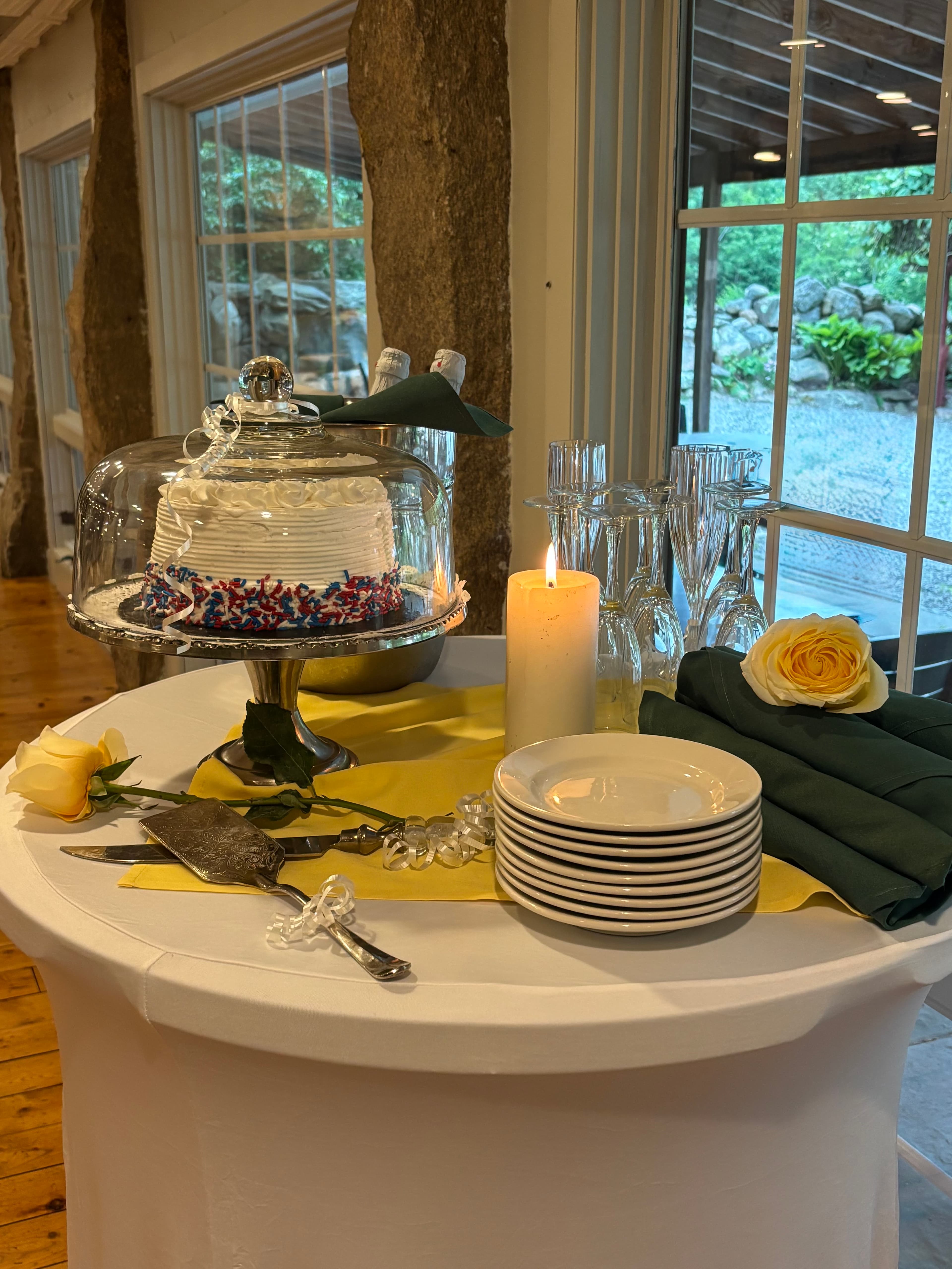 A decorated cake under a glass dome, surrounded by a candle, glassware, plates, and yellow roses on a table.