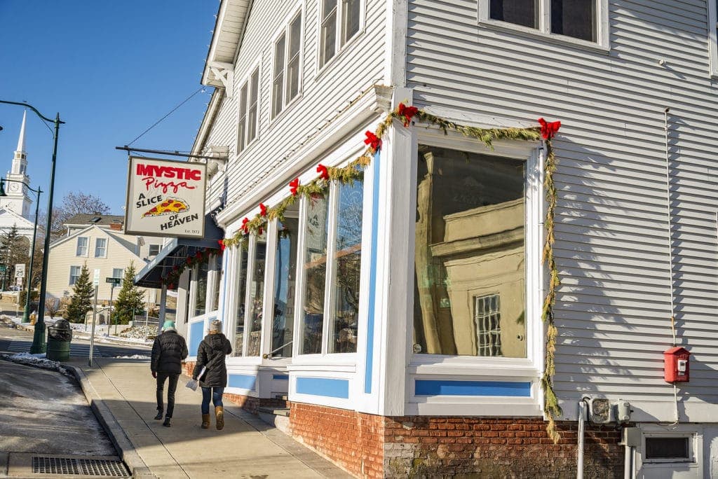 A couple walks past the decorated exterior of Mystic Pizza on a sunny day.