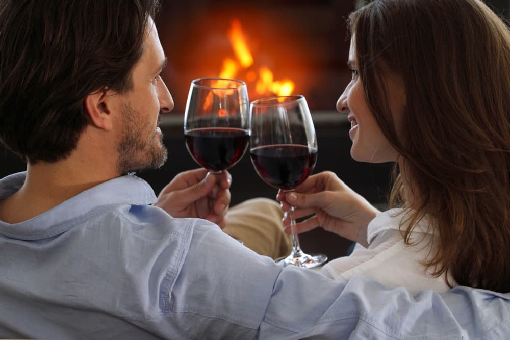 A couple toasts with glasses of red wine in front of a cozy fireplace.