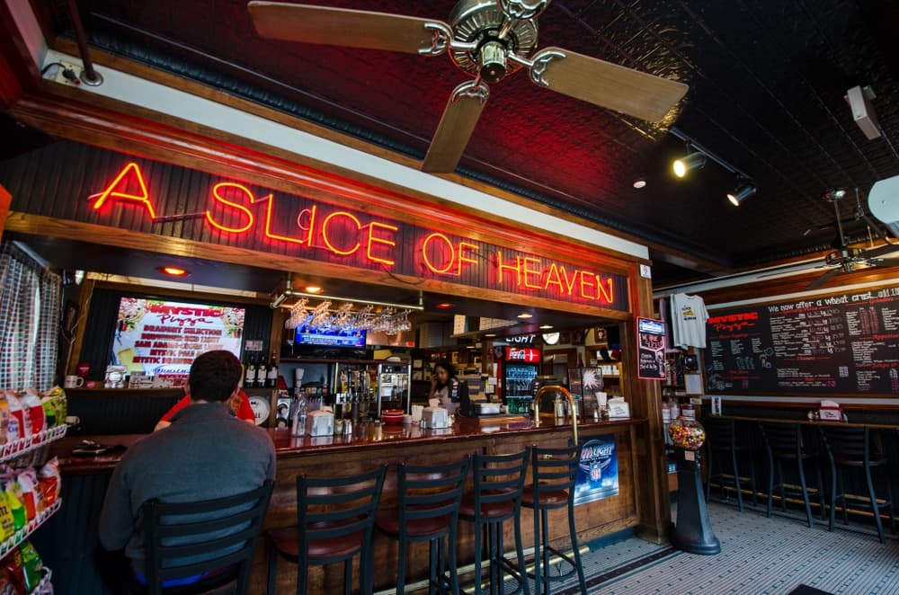 Interior of a bar with a neon sign reading "A Slice of Heaven" and a patron seated at the counter.