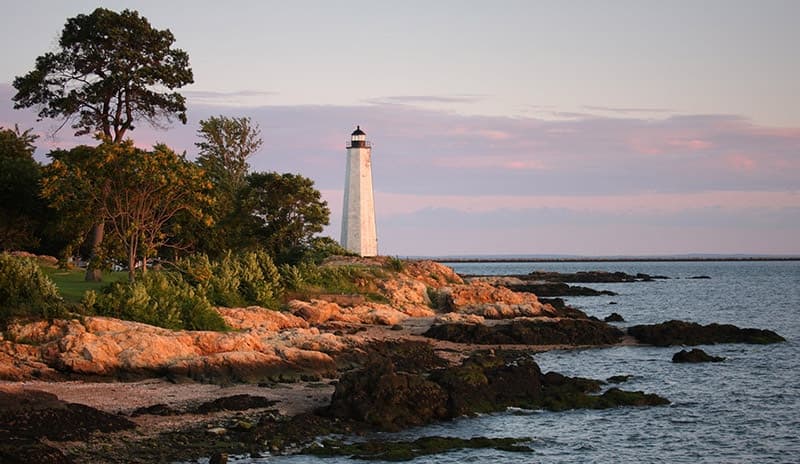 A lighthouse stands on rocky shorelines under a pastel sunset sky.