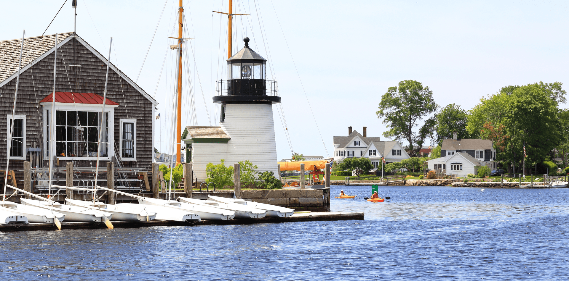A lighthouse stands beside a marina with sailboats, framed by a calm waterfront and residential homes.