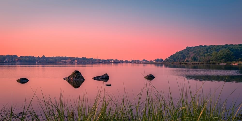 A tranquil lake at sunset, reflecting soft pink hues and surrounded by greenery and rocks.