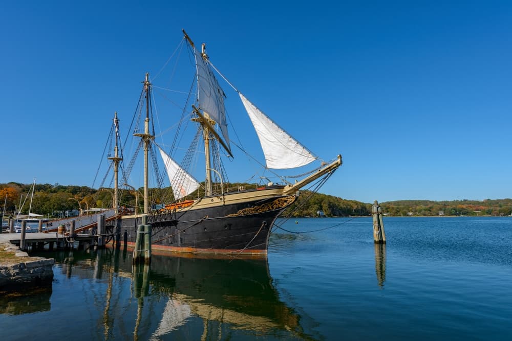 A tall ship with sails raised is docked by a calm, reflective body of water under a clear blue sky.
