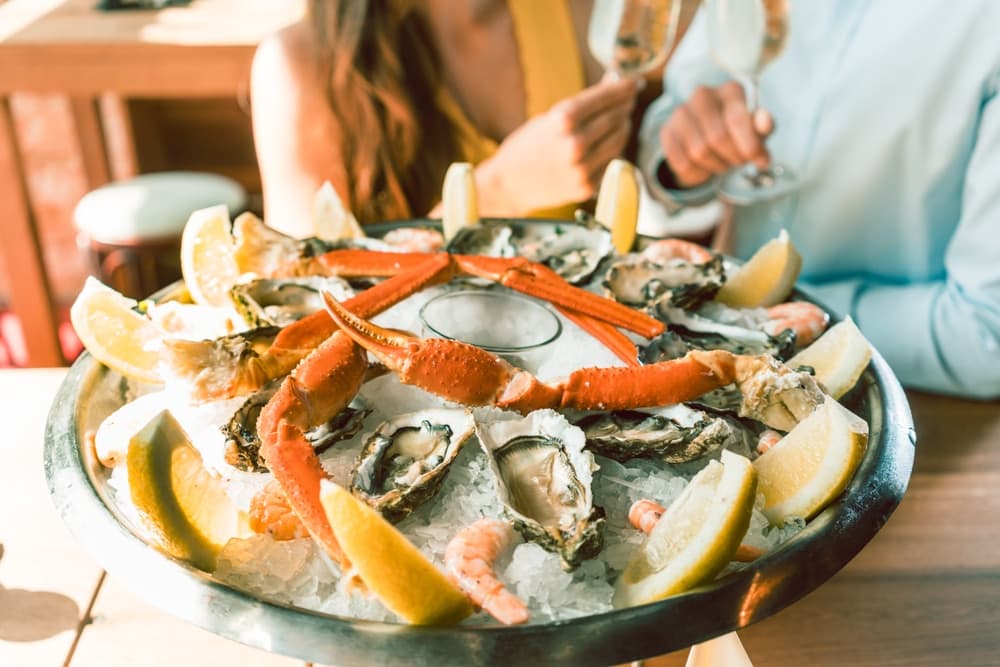 A seafood platter with oysters, crab legs, shrimp, and lemon wedges on ice, accompanied by two people enjoying drinks.