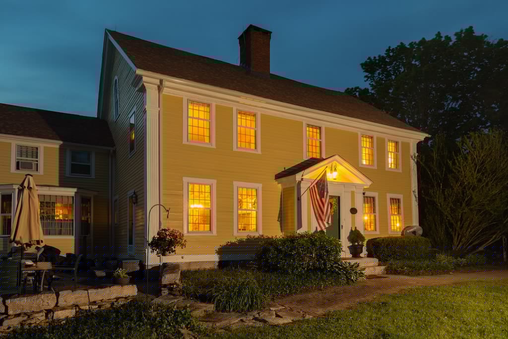 A charming yellow house illuminated at dusk, featuring glowing windows and an American flag.