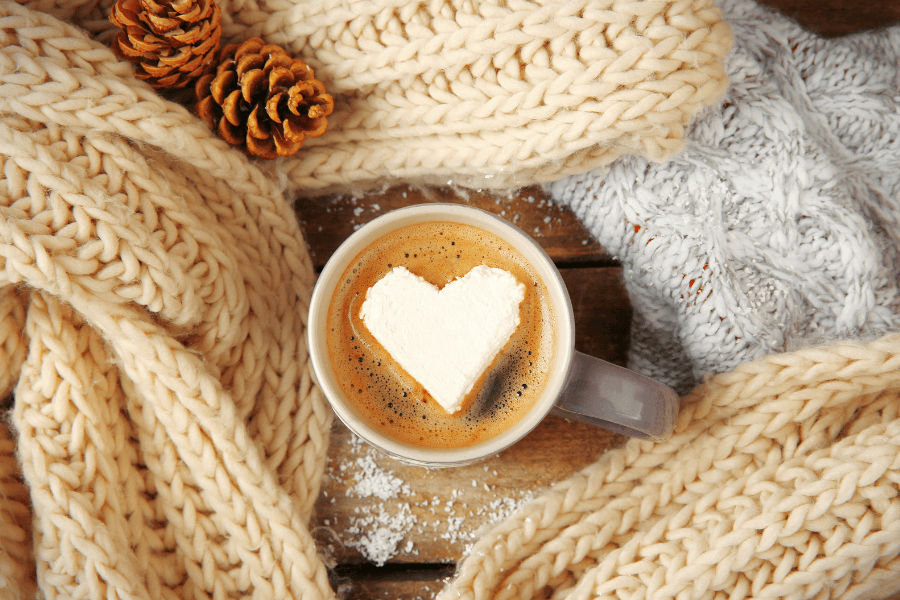 A cozy cup of coffee topped with a heart-shaped dollop of cream, surrounded by soft knitted scarves and pinecones.