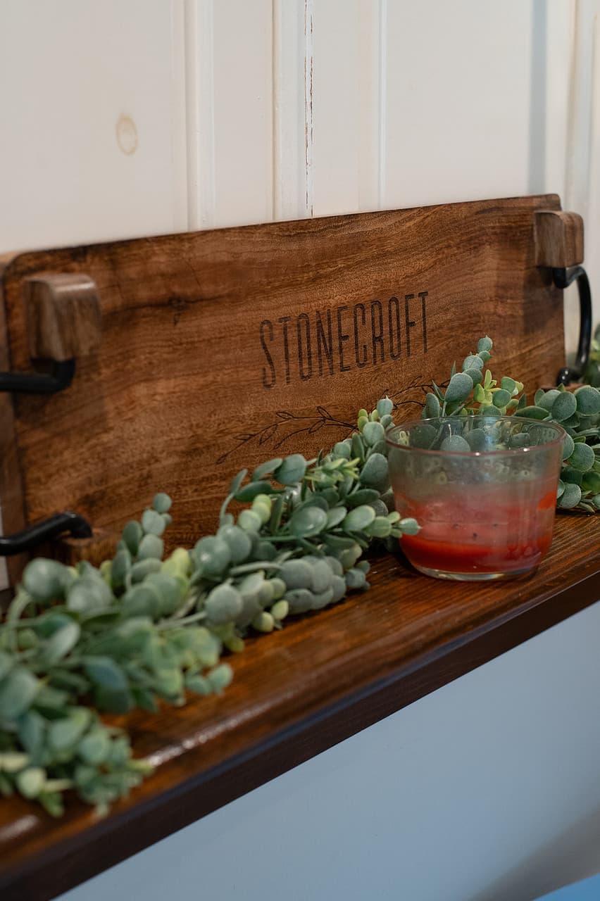A wooden sign labeled "STONECROFT" rests on a shelf adorned with green foliage, alongside a small glass bowl containing red liquid.