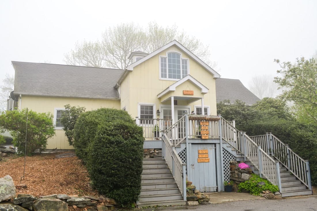 A quaint yellow house with a blue door and wooden steps, surrounded by greenery and shrouded in fog.