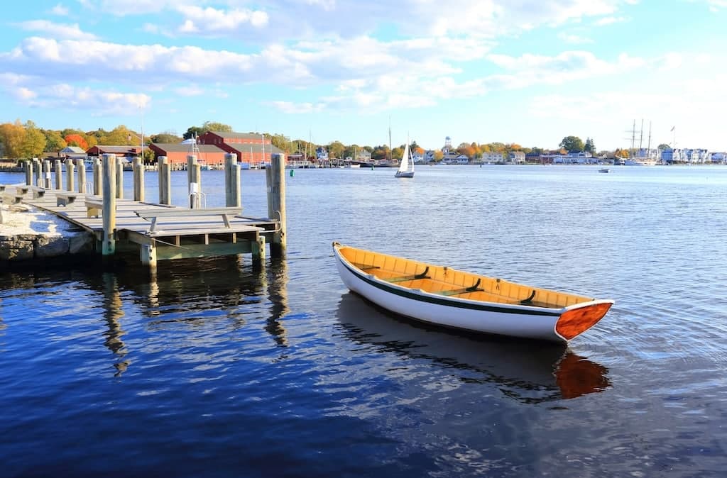 A wooden boat is moored near a pier on a calm body of water, with colorful trees and buildings in the background.