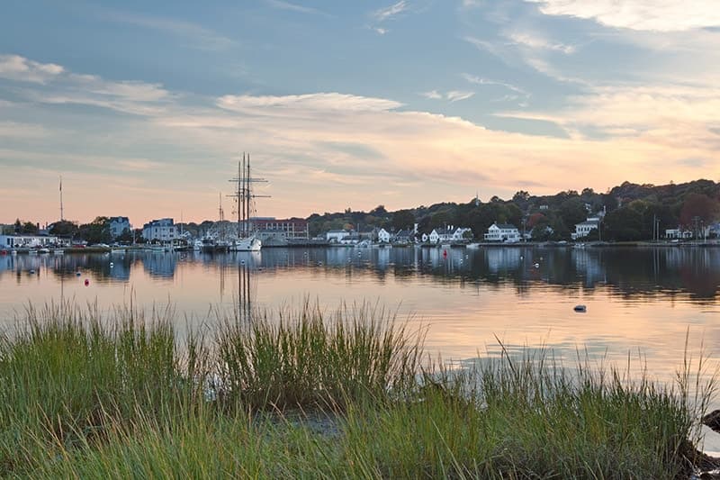 A tranquil waterfront scene with grass in the foreground and a small village reflected in calm waters at sunset.