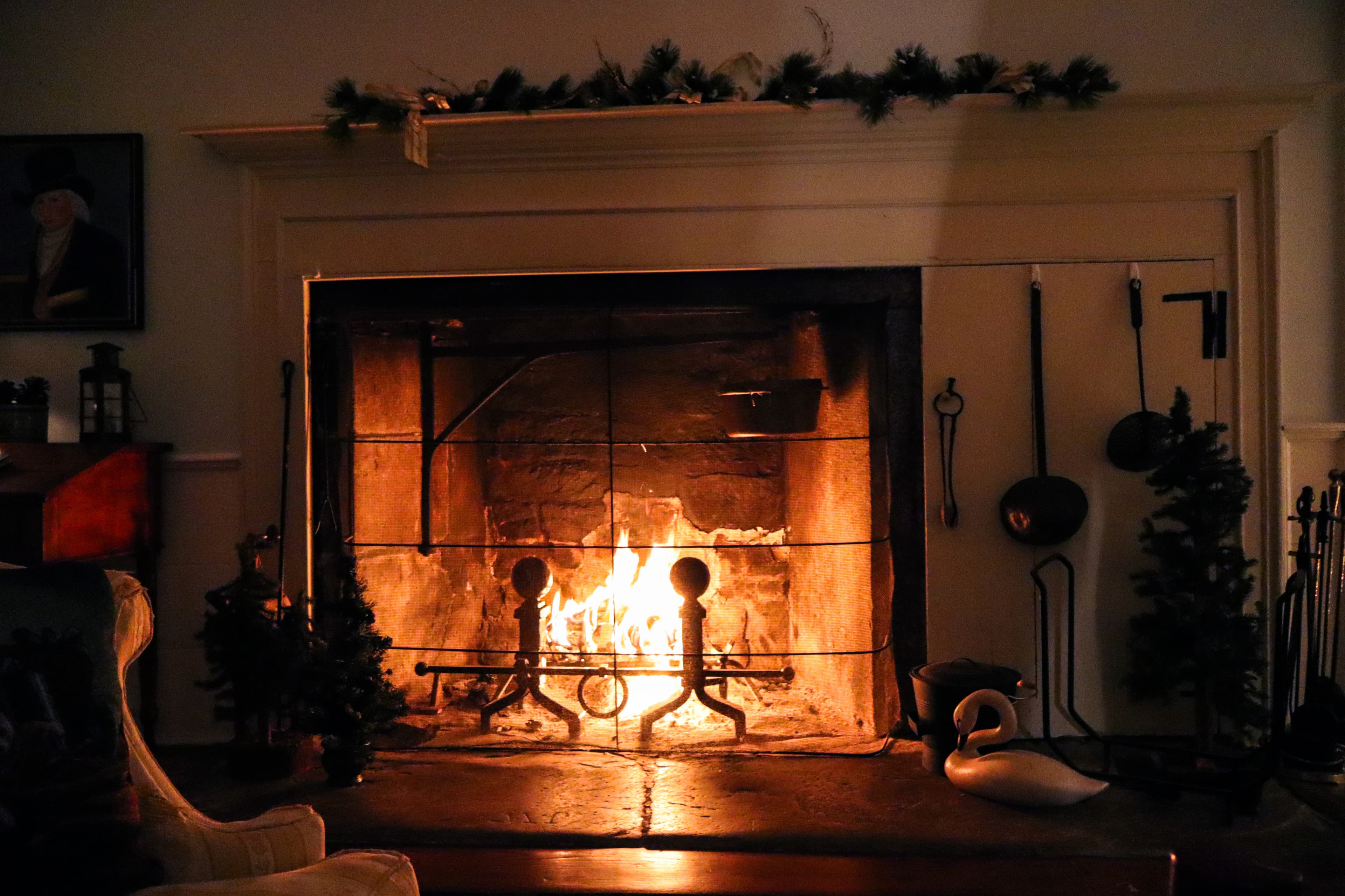 A warmly lit fireplace adorned with festive greenery and tools, casting a cozy glow in a dimly lit room.
